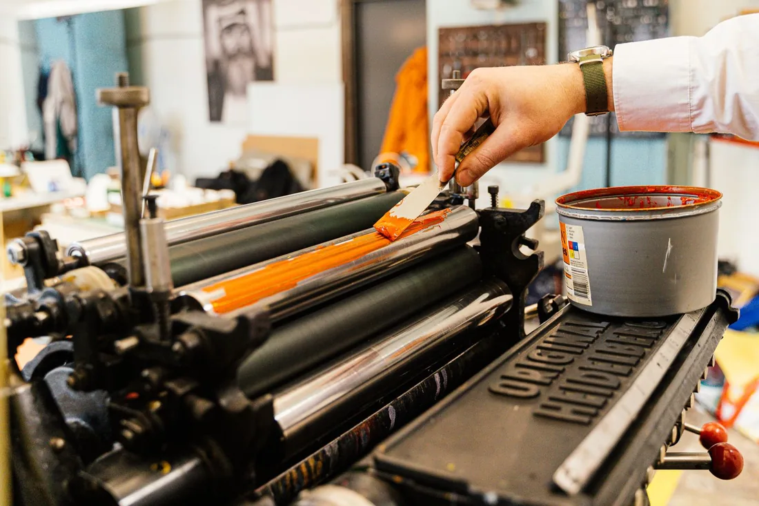 Mano aplicando tinta anaranjada sobre el rodillo de una máquina de impresión en un taller